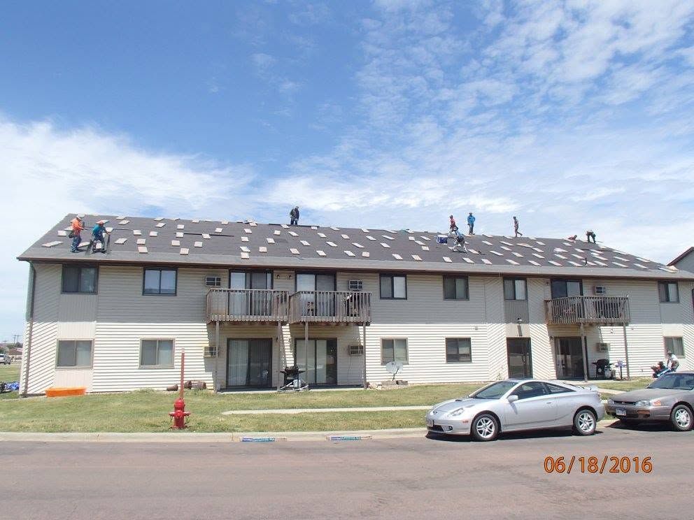 Two-story apartment building with workers on the roof and a silver car parked in front.