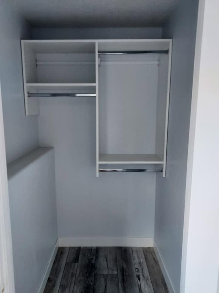 Small empty walk-in closet with white shelves and hanging rods, gray walls, and wood-look flooring.