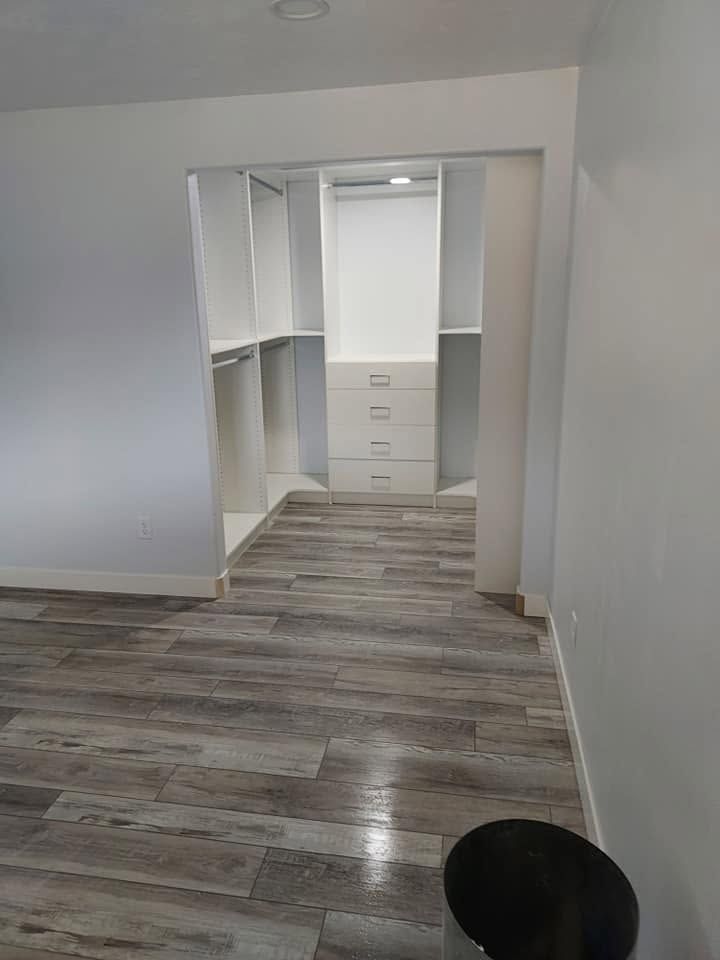 Empty closet with white shelving and drawers, gray wood-look flooring, and a black trash bin in the foreground