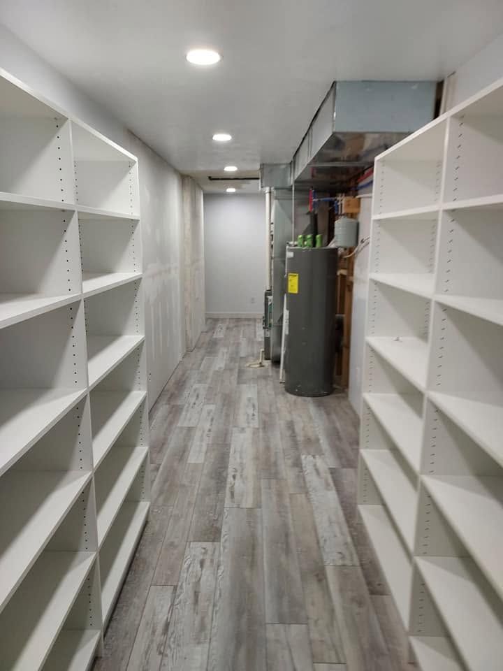 Narrow storage hallway with white shelves, gray wood floor, ceiling lights, and a water heater at the end.