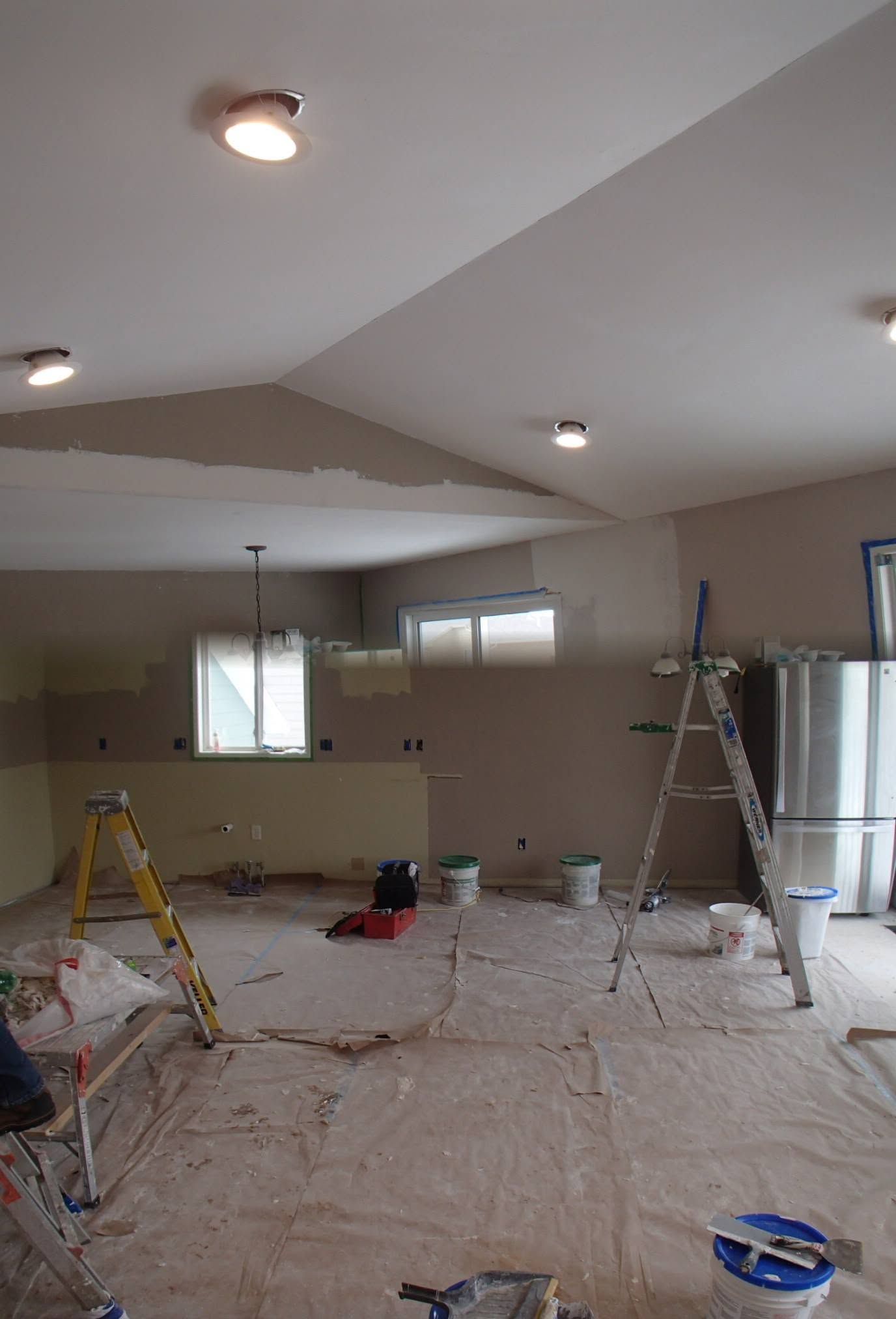 Partially renovated room with covered floor, ladders, paint supplies, and ceiling lights on