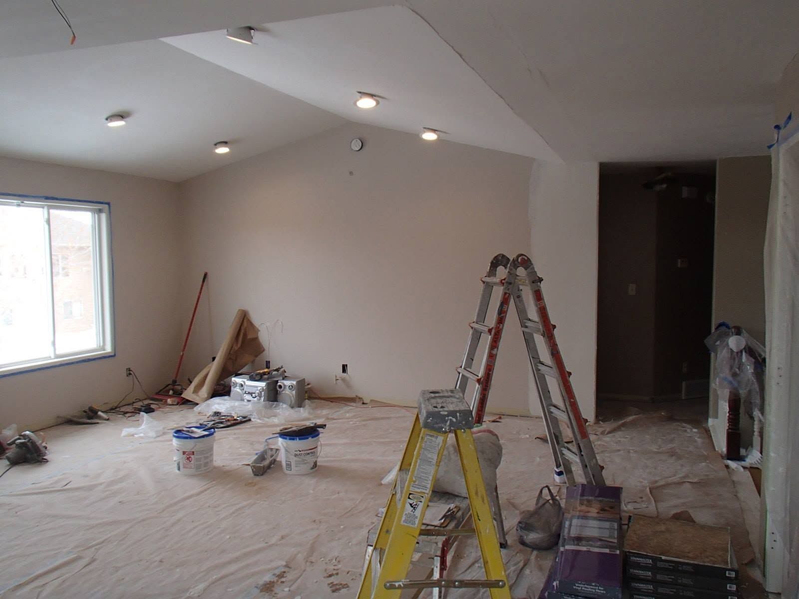 Partially renovated room with ladders, paint buckets, and plastic-covered floor under a sloped ceiling