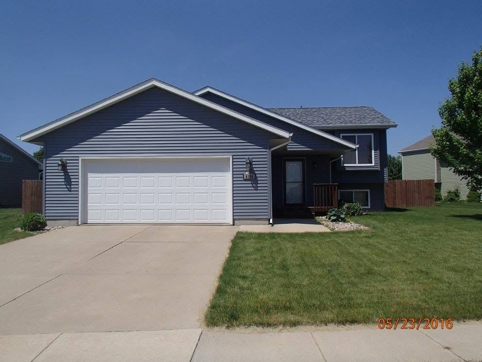 Single-story gray house with white garage door, concrete driveway, and a front lawn under a clear blue sky