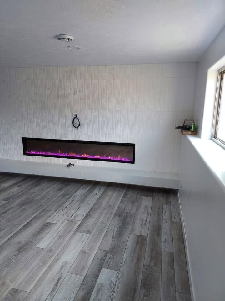 Empty modern living room with gray wood floors, white walls, and a long black electric fireplace with pink flames.