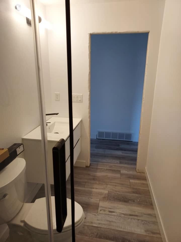 Narrow hallway with a toilet on the left, leading to a blue-walled room with wood-look flooring.