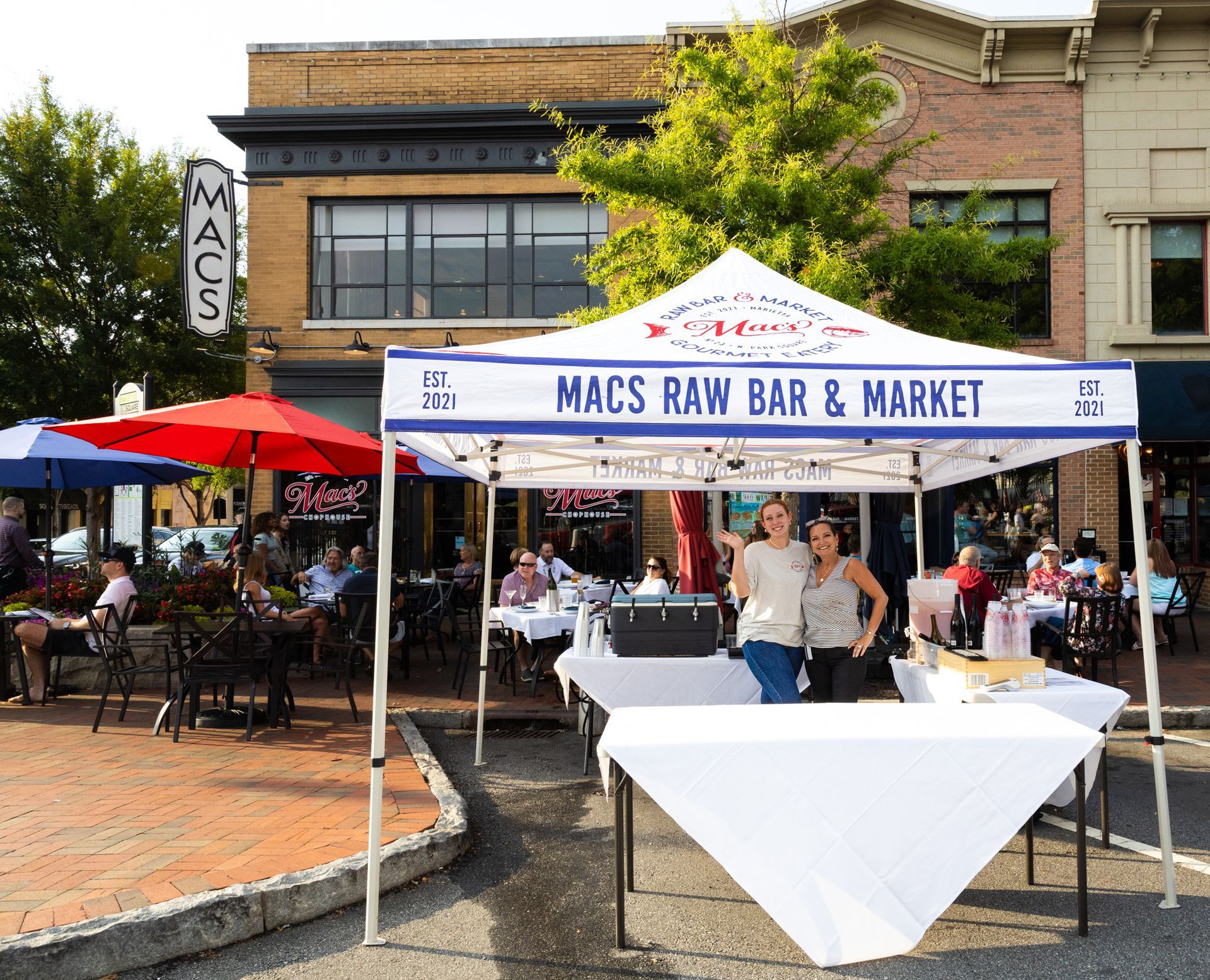Two women standing under a macs raw bar and market tent