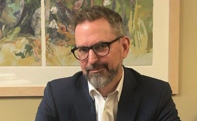 Man in glasses and navy suit, hands clasped, looking at camera. Painting in background.