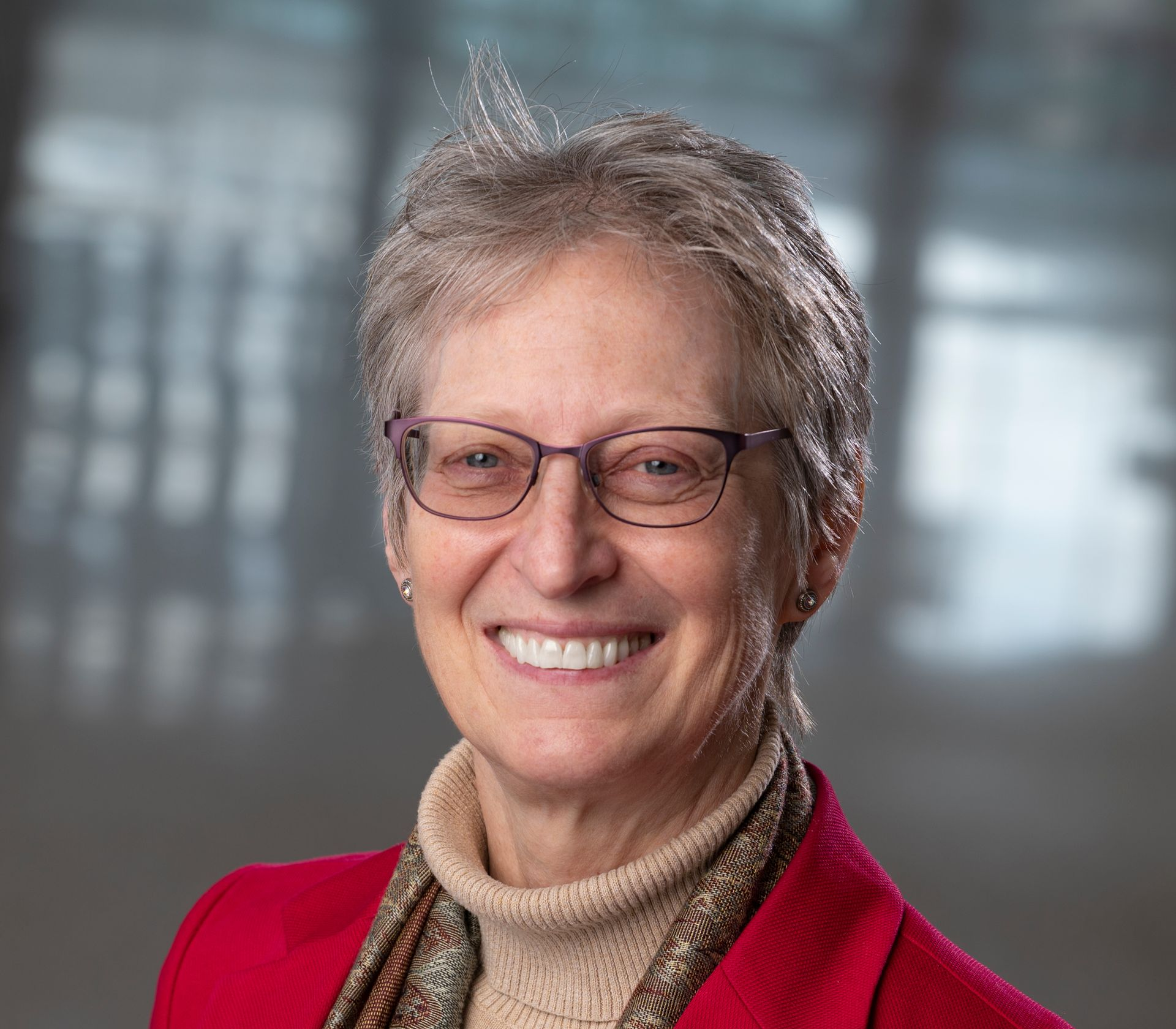 Woman with short gray hair, wearing glasses, a red jacket, smiling in a professional setting.