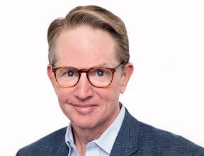 A man with light brown hair wearing glasses and a blue patterned blazer against a white background.