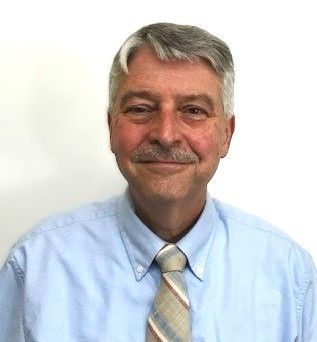 A man with gray hair and a mustache smiles while wearing a light blue button-down shirt and a patterned necktie.