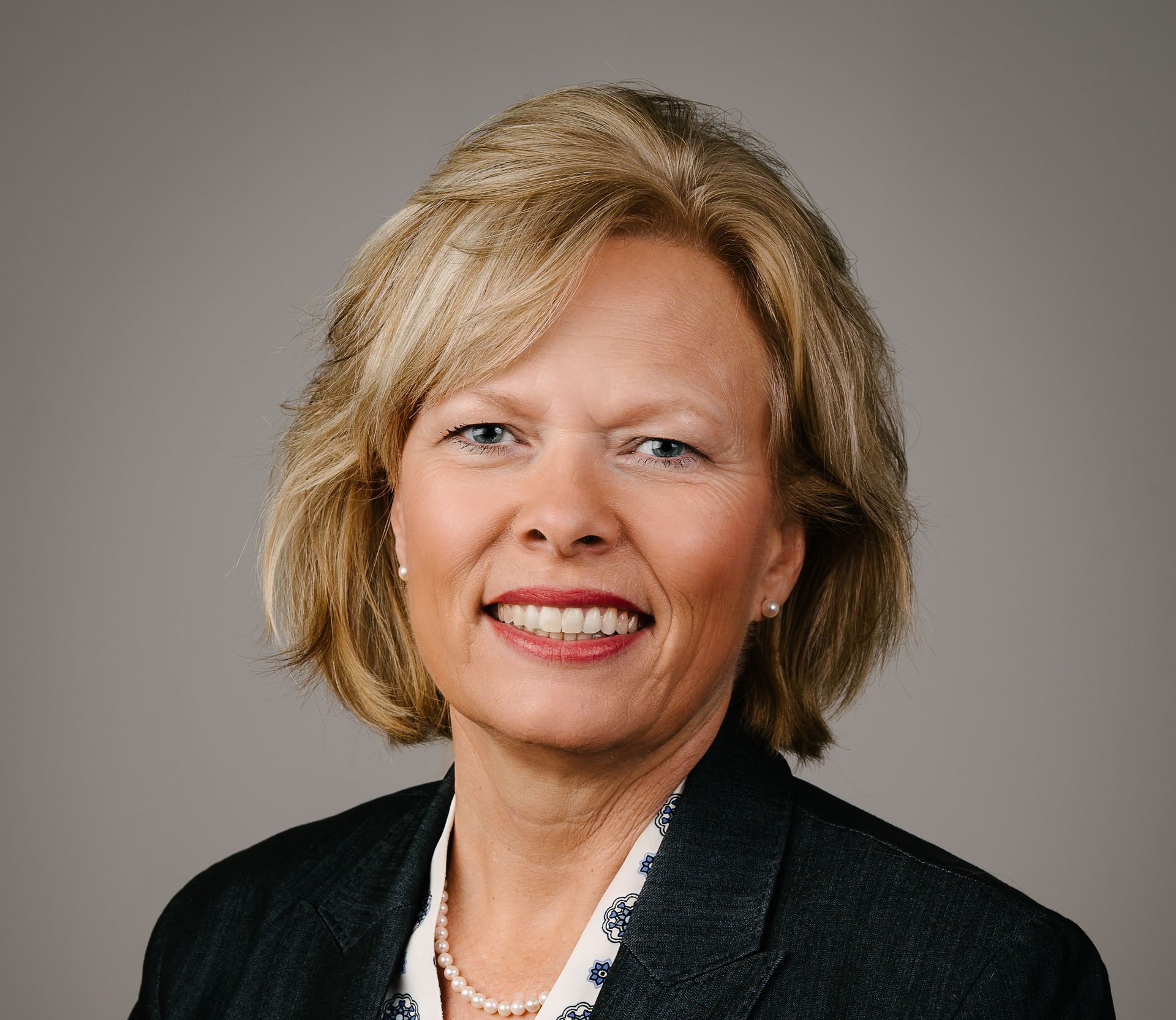 Woman in blazer smiles, posed headshot against a gray backdrop.