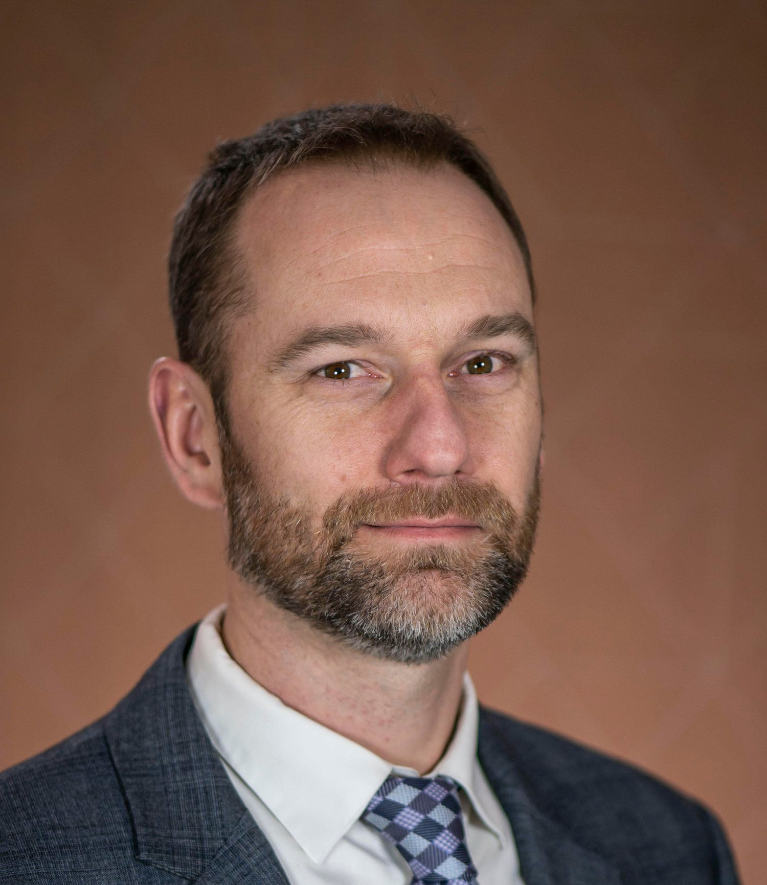 Man in suit and tie looking directly at the camera, neutral expression, against a brown background.