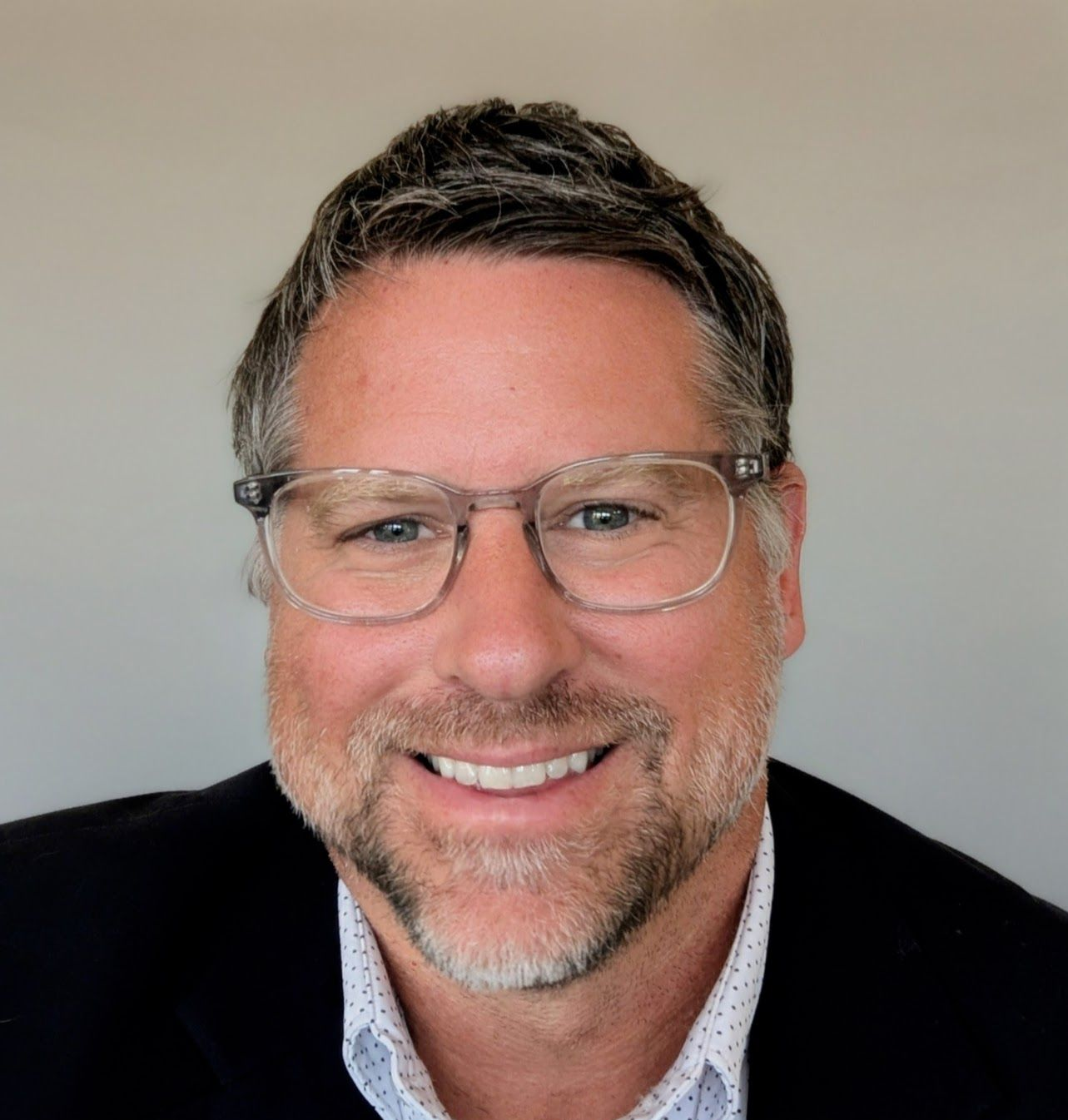 Man wearing glasses smiles, in a blazer over a button-down shirt.