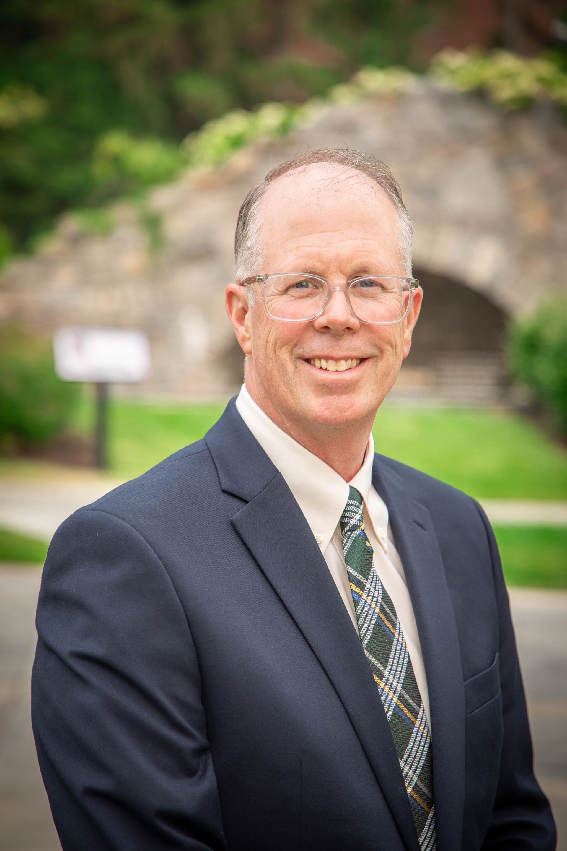 Smiling man in a navy suit and patterned tie standing outdoors with a blurred stone archway behind him