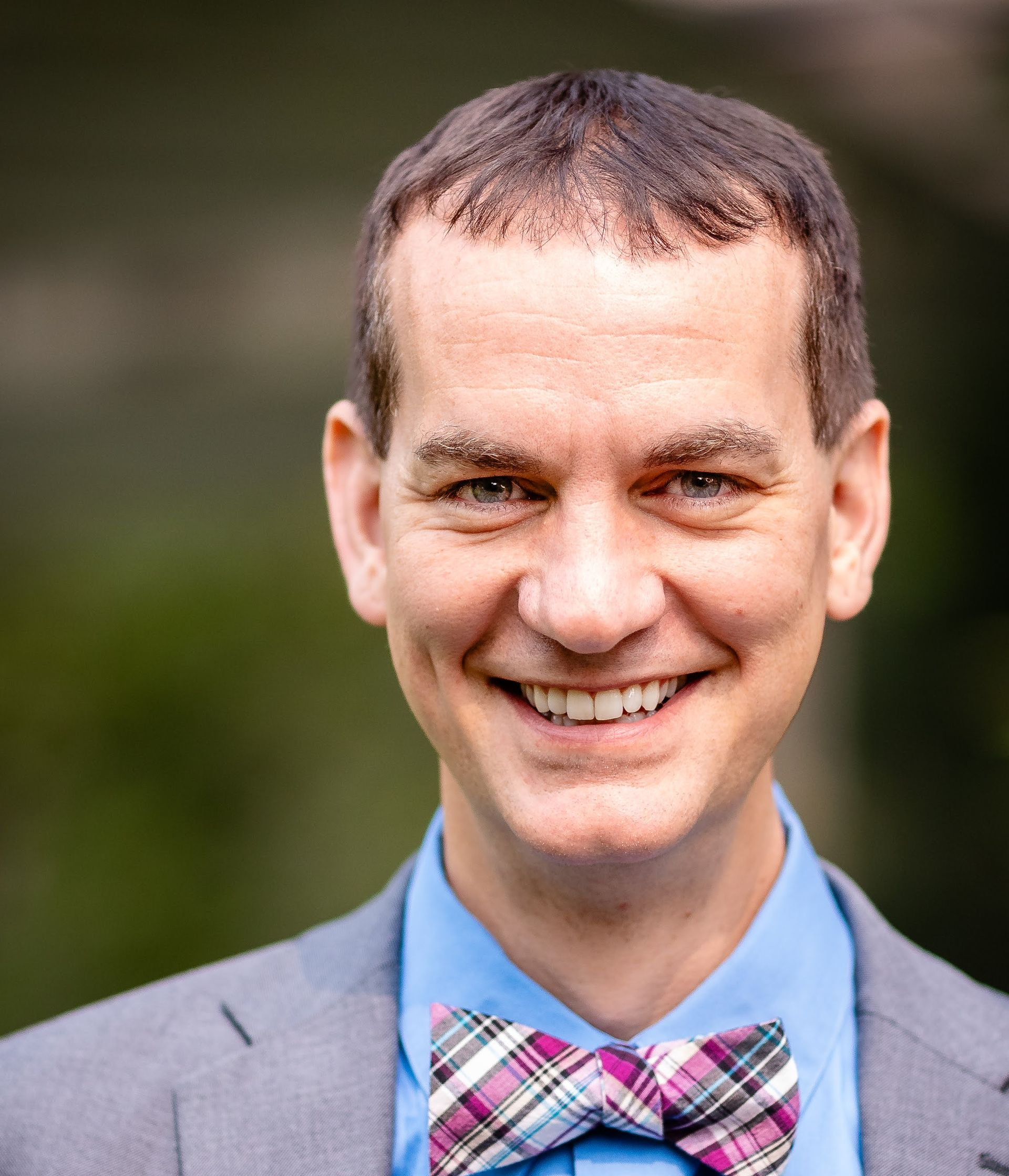 Man wearing a blue shirt, gray suit jacket, and plaid bow tie, smiling broadly outdoors.