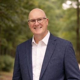 Man with a beard and graying hair, smiling, wearing a blue blazer and button-down shirt, in front of greenery.
