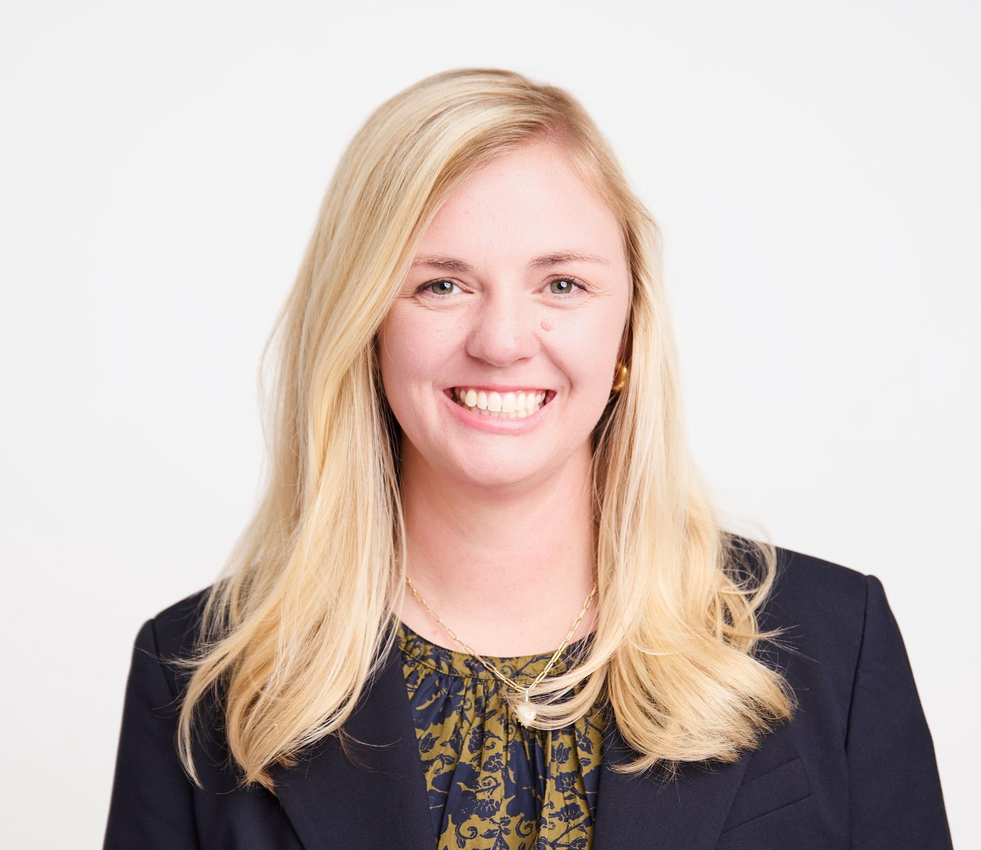 A smiling blonde individual with a gold necklace, wearing a dark navy blazer over a patterned dark-green top.