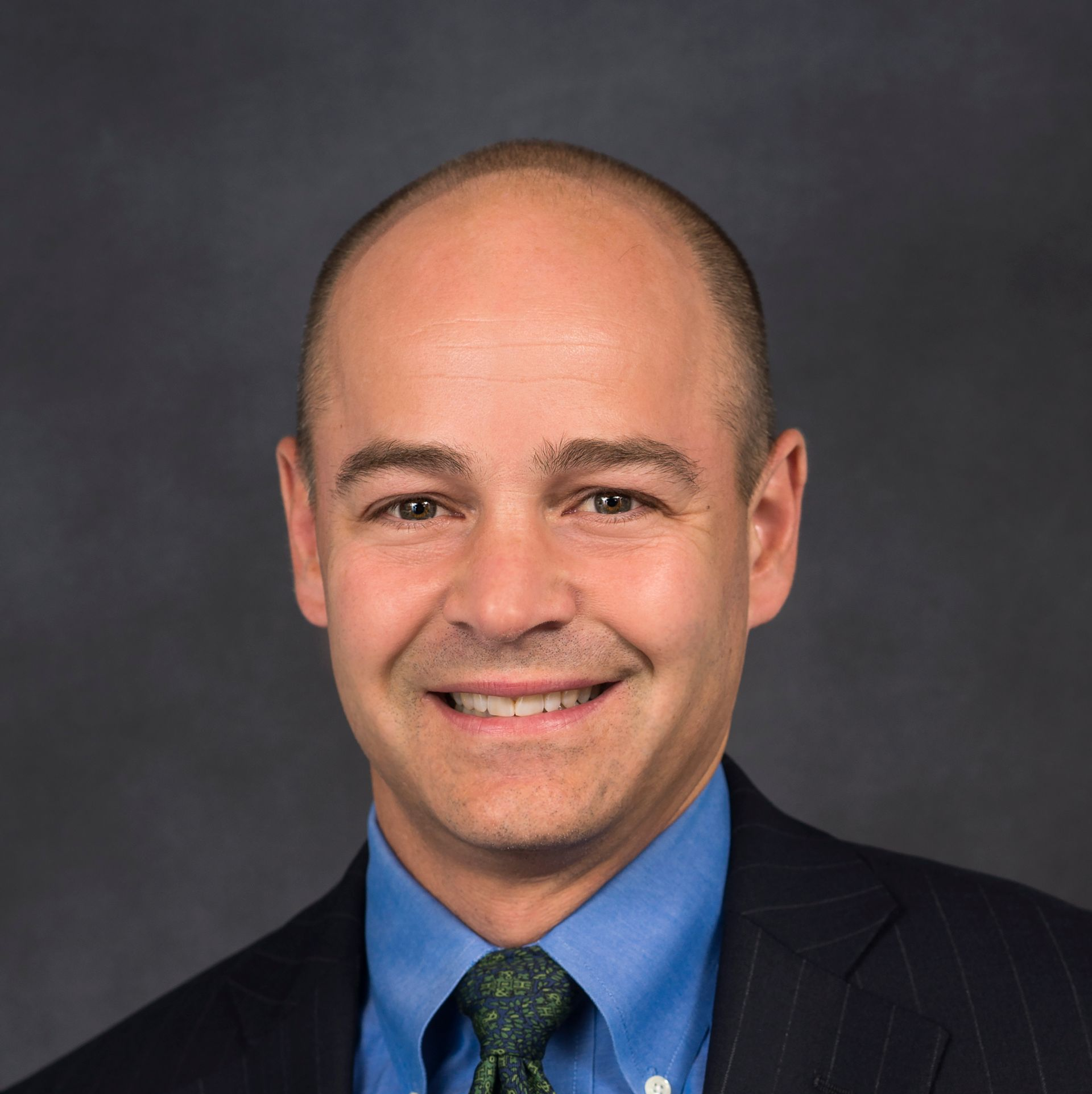 Man in blue shirt, green tie, and suit jacket smiles at the camera against a gray backdrop.