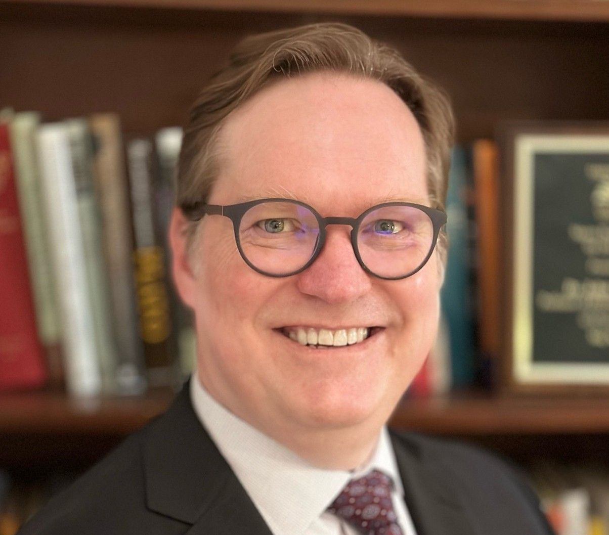 Smiling professional in a dark suit, light shirt, and patterned tie, posing in front of a bookshelf.