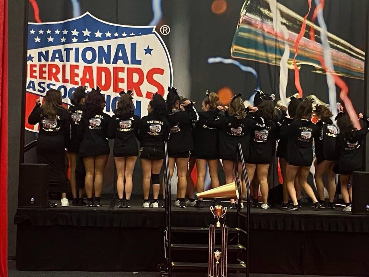 A cheerleading team in black uniforms stands on a stage in front of a National Cheerleaders Association banner.