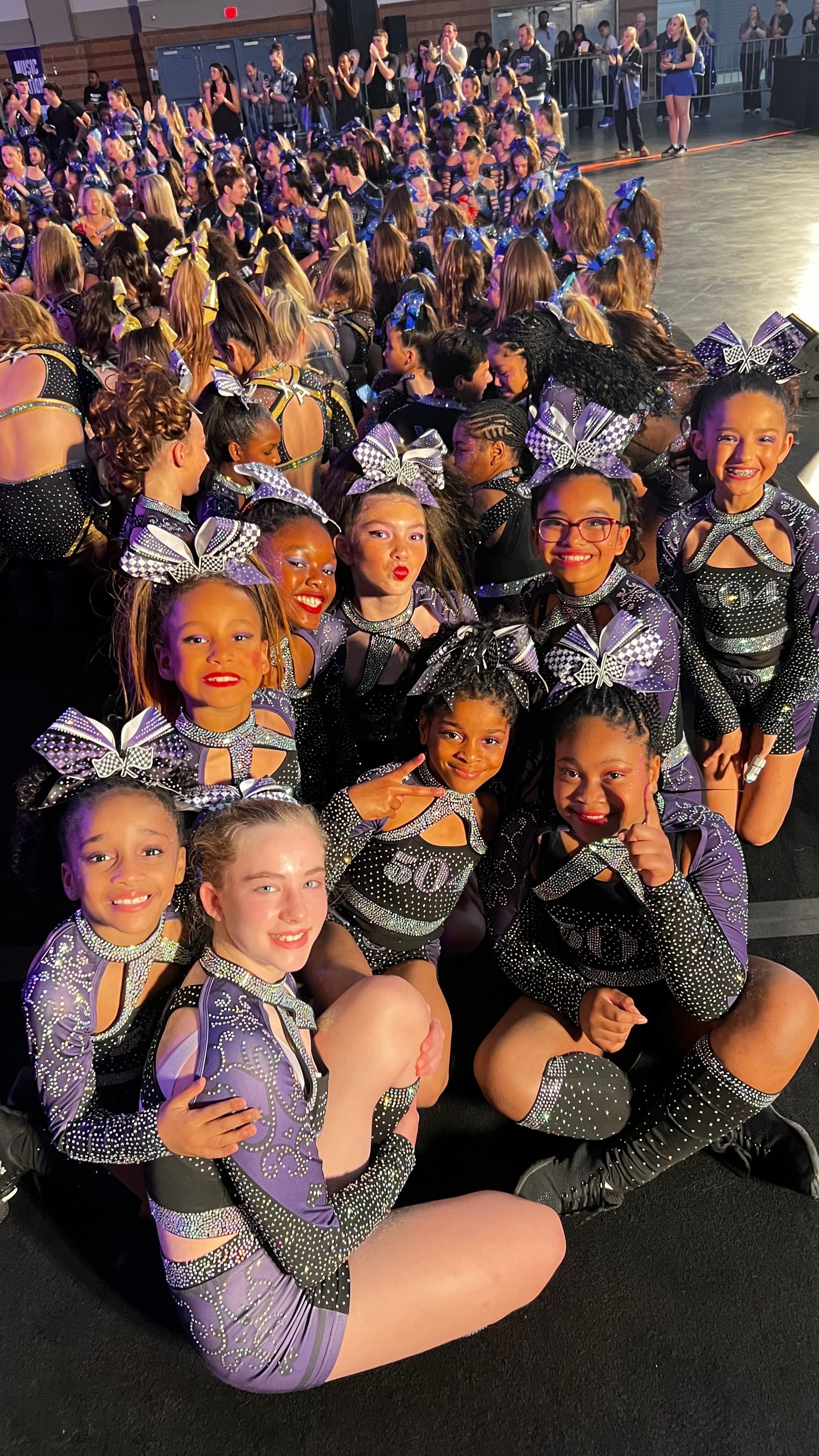A group of cheerleaders in matching purple and black sequined uniforms posing together at an indoor competition venue.