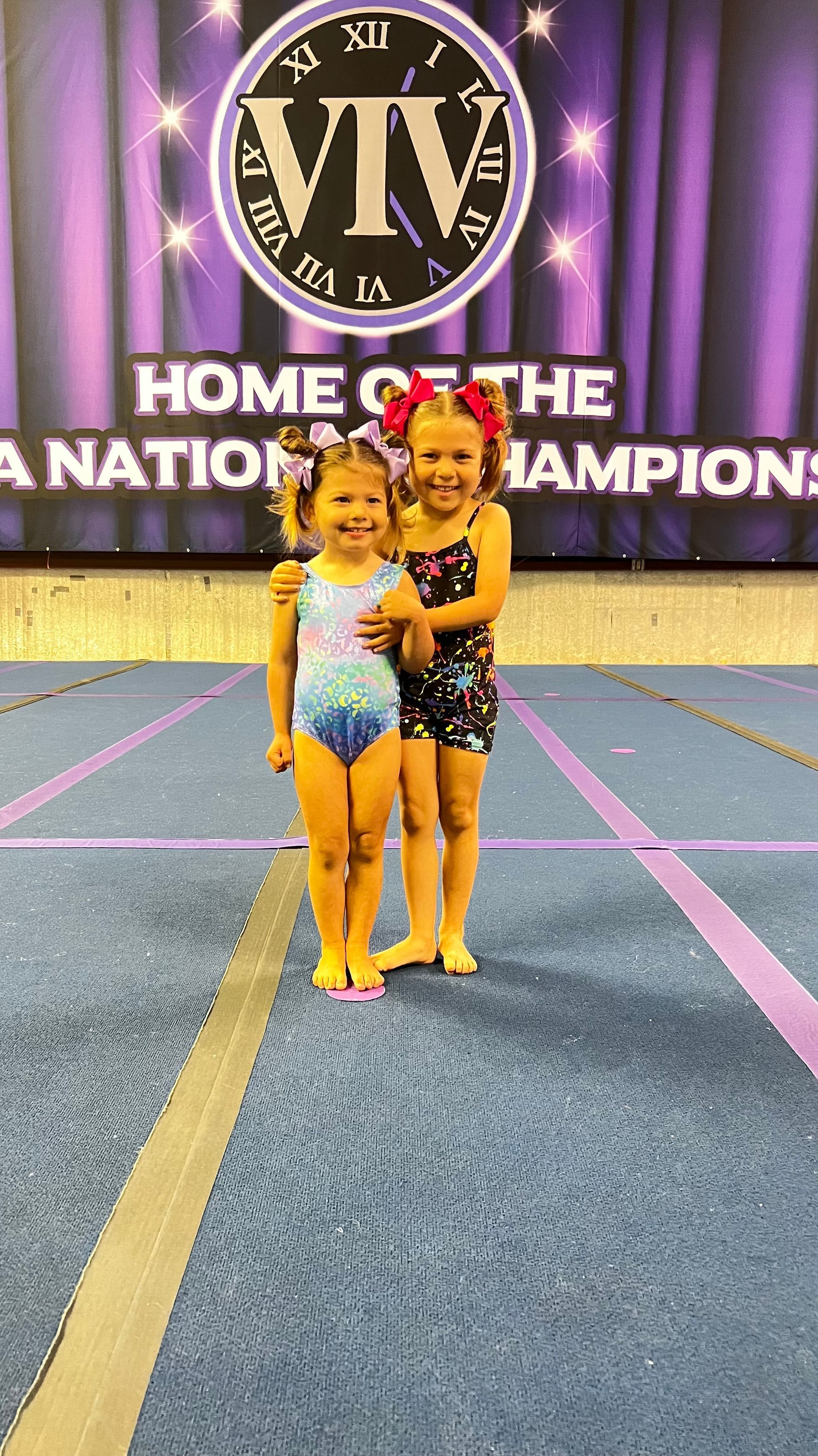 A cheerleader in a purple and black uniform posing on a blue competition mat in front of a banner.