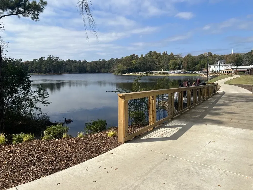 A concrete path follows a wooden railing along a calm lake, with trees lining the opposite shore under a blue sky at The Reserve at Mill Landing.