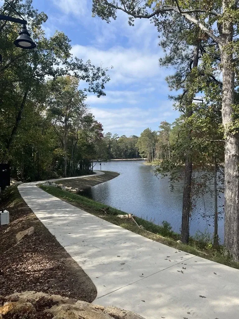 A paved walking path winds along the edge of a calm lake surrounded by trees under a blue sky with light cloud sat The Reserve at Mill Landing.