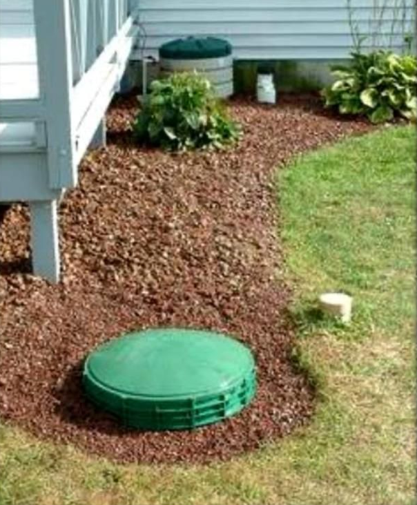 Green septic tank lid in mulch, next to a deck and lawn. Additional septic tank and plants visible.