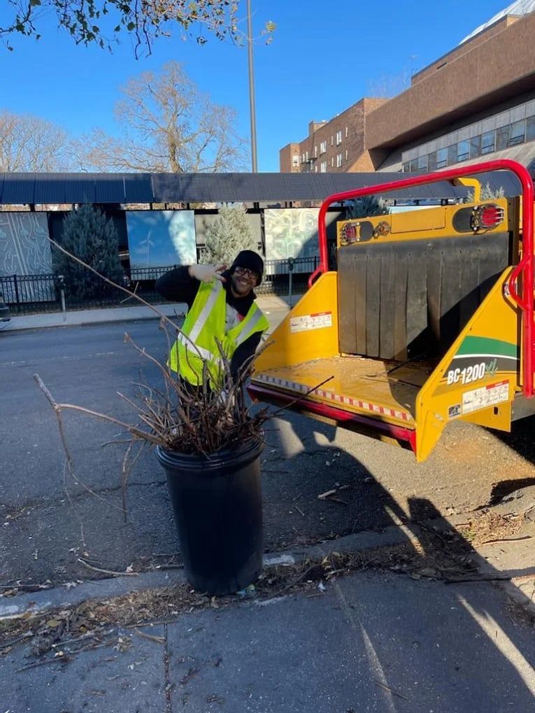 Worker in a neon safety vest beside a yellow utility trailer and potted plant on a street.