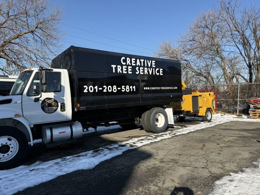 Tree service truck with trailer and yellow equipment parked beside a snowy roadside.