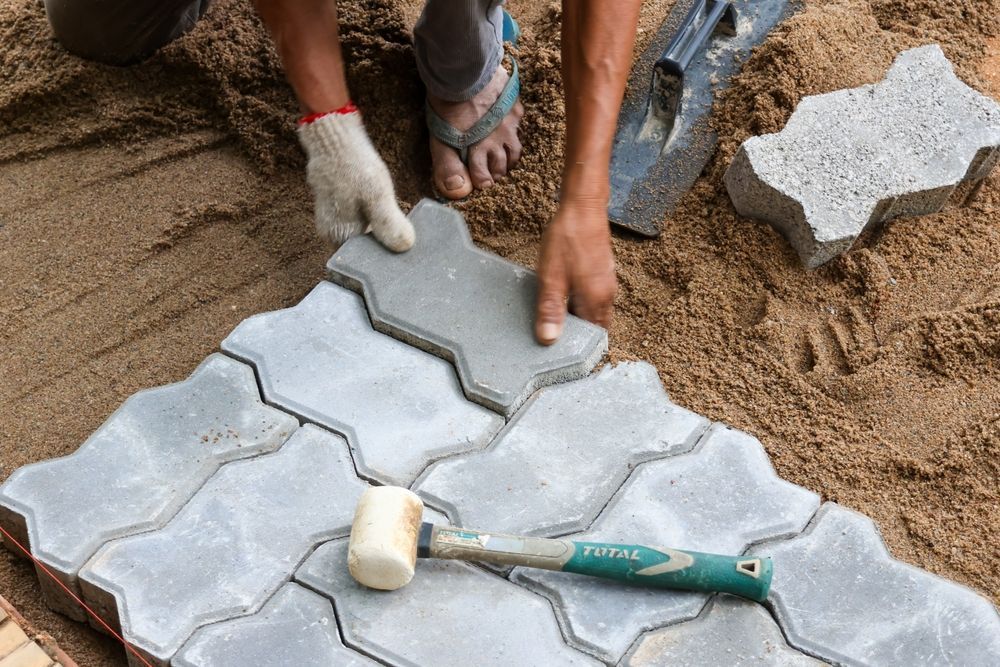 Hands laying gray interlocking pavers on sand with a rubber mallet nearby