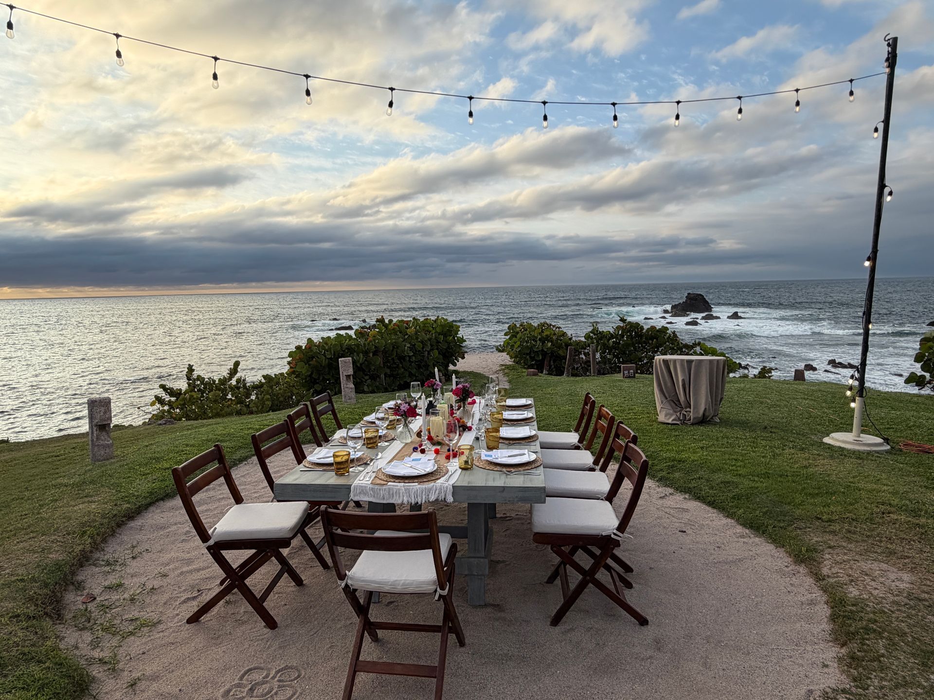 Outdoor dining table set on a grassy seaside cliff at sunset, overlooking the ocean.