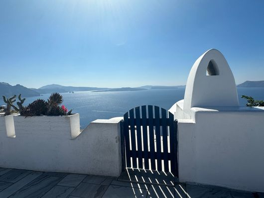 White gate overlooking a bright blue sea and sky, with a sunlit white wall and archway.