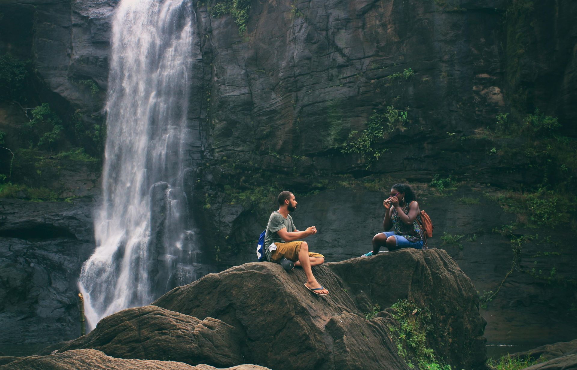 Two people sitting on a large rock, talking in front of a tall, thin waterfall in a wooded, rocky setting.