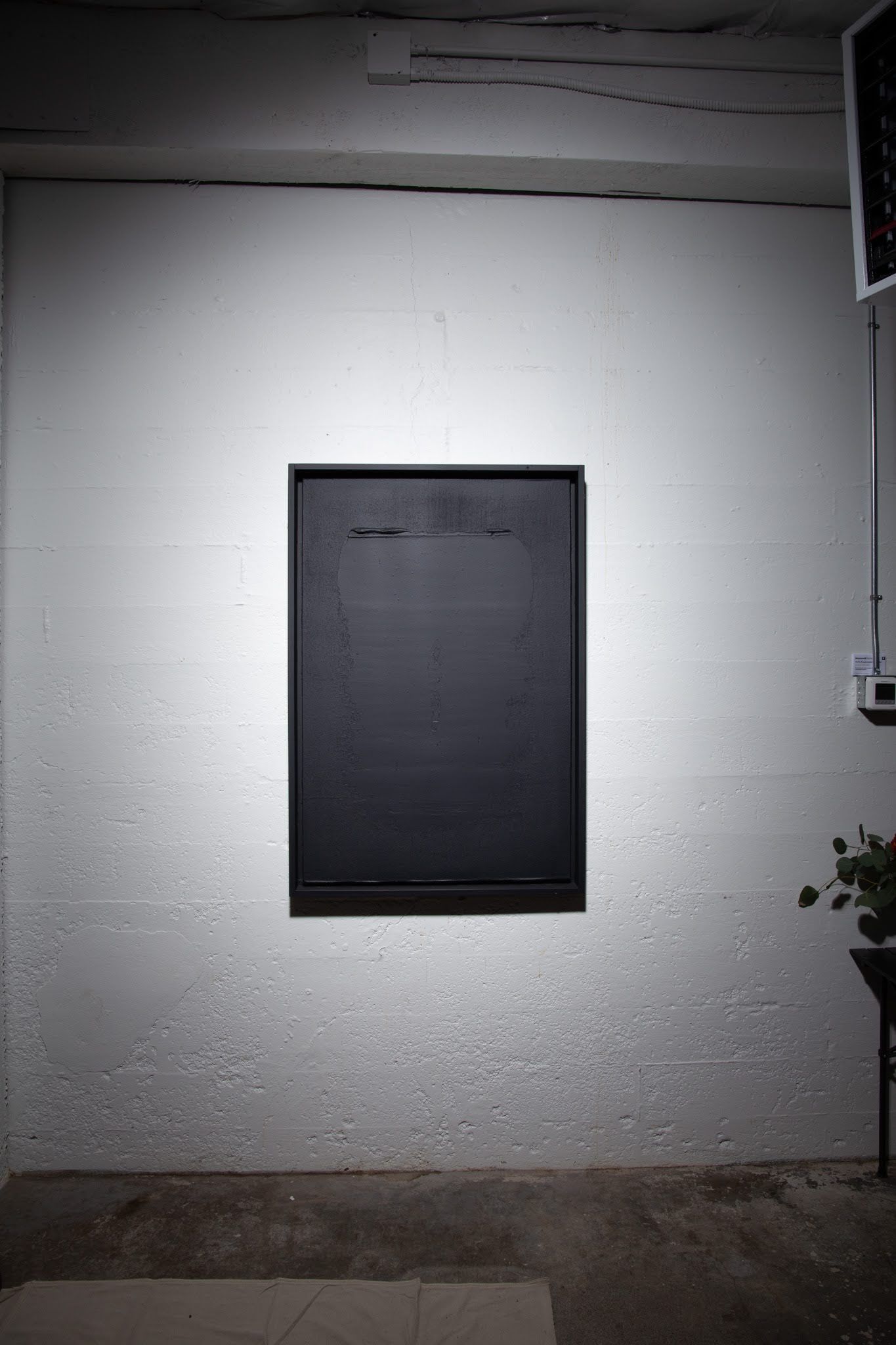 Black rectangular panel on a textured white wall, spotlighted in a dimly lit room; art by Matt Remsbecher. 
