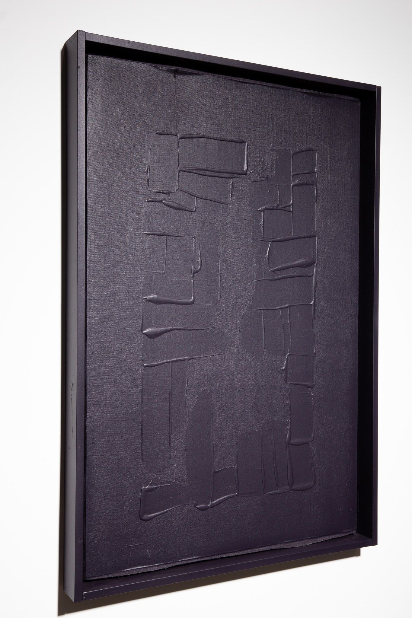 Dark textured rectangular abstract art in a black frame, leaning against a white wall; art by Matt Remsbecher. 