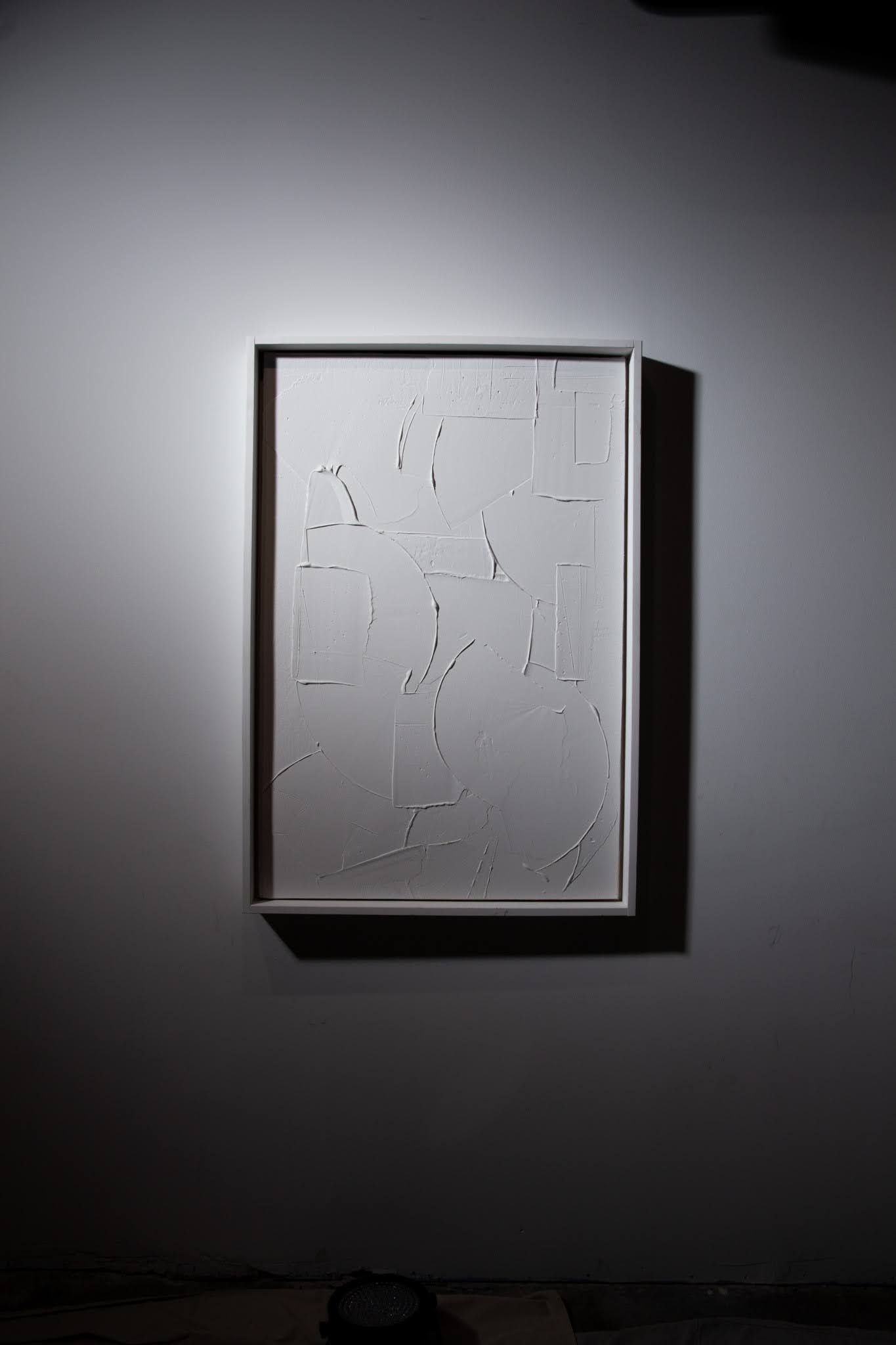 White framed art piece by Matt Remsbecher on a light gray wall. The art is white with scattered shapes, under a spotlight.