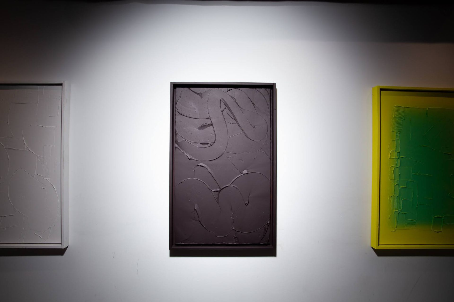 Three abstract paintings by Matt Remsbecher on a white wall. Dark textured art is between a white and a green artwork.