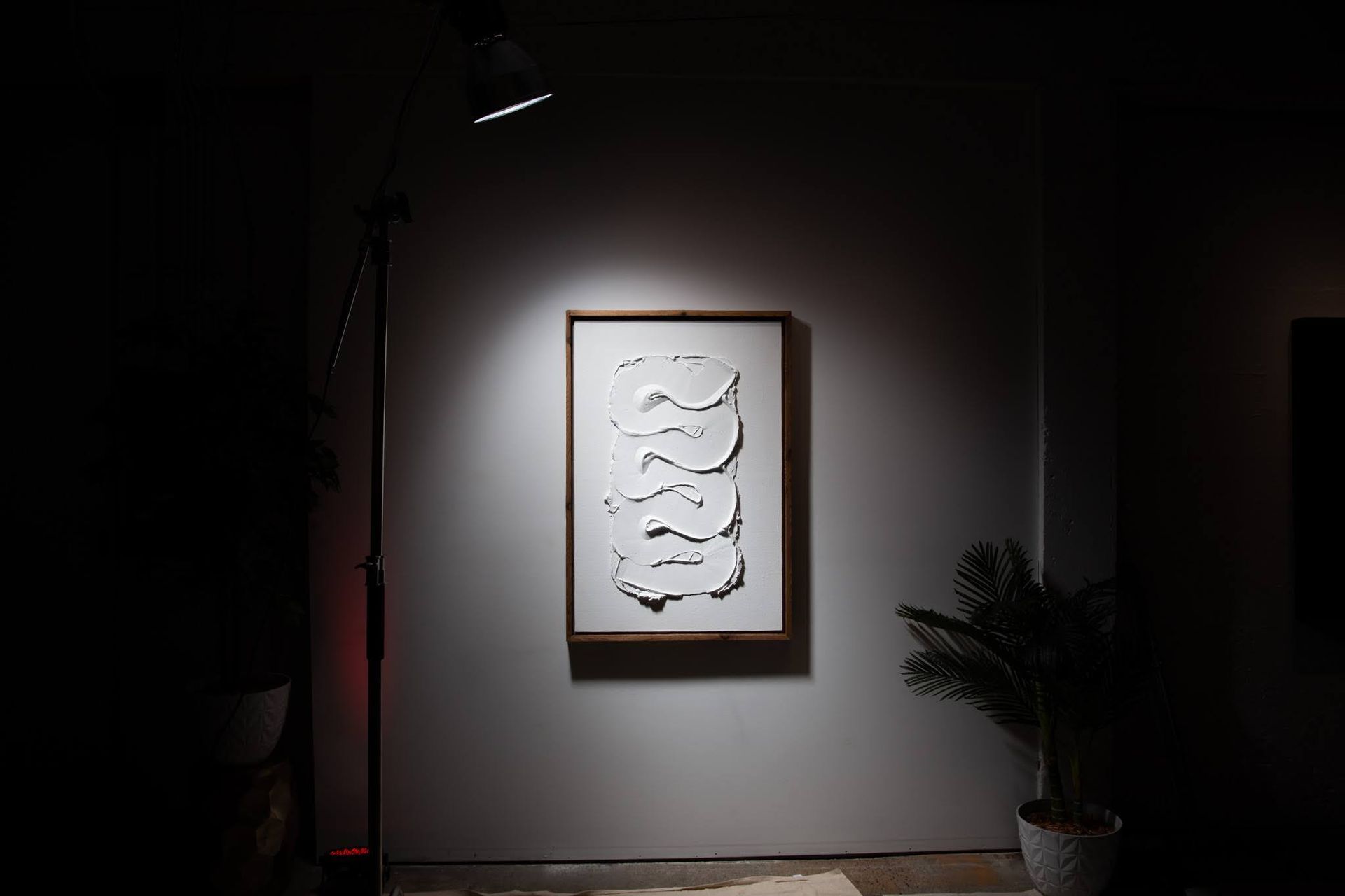 A spotlight illuminates an abstract white relief sculpture in a wooden frame on a gray wall; art by Matt Remsbecher. 