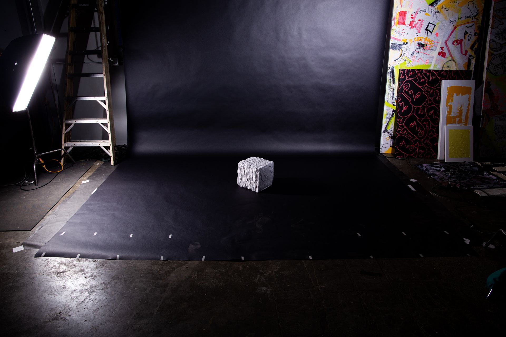A cube sits on a black backdrop in a studio setting, lit by a side light and a ladder is visible; art by Matt Remsbecher. 