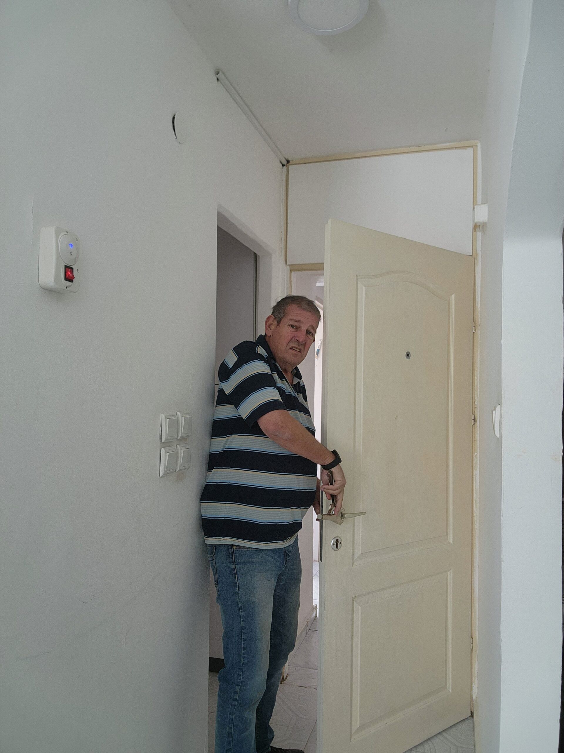 מנעולן 24/7 בחולצת פסים עומד בפתח דלת פנים לבנה ופתוחה בתוך מסדרון של בית ומחזיק בידית הדלת.