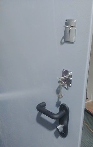 תקריב של דלת פלדה אפורה שעליה מותקנים שני מנעולי כספת עליונים עם מפתח תקוע באחד מהם מעל ידית דלת שחורה.
