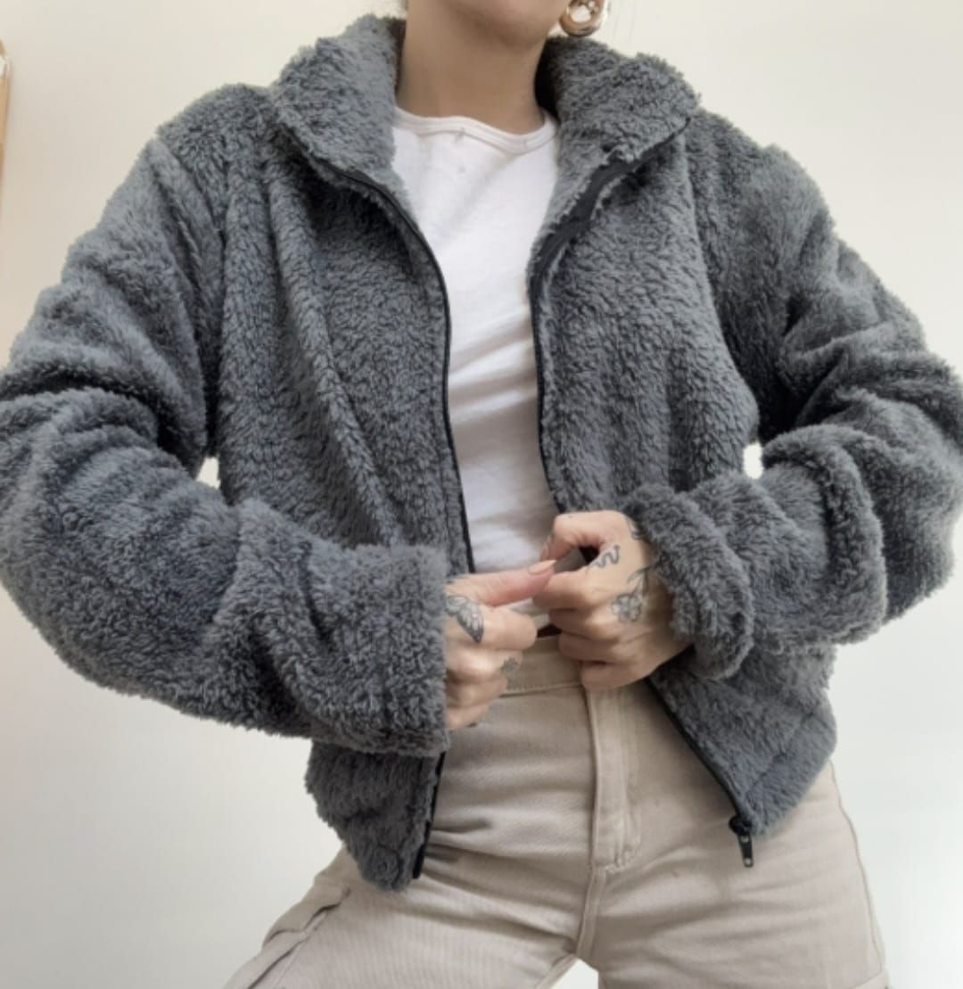 Persona que lleva una chaqueta gris de pelo sintético sobre una camisa blanca, ajustándose la cremallera delantera.