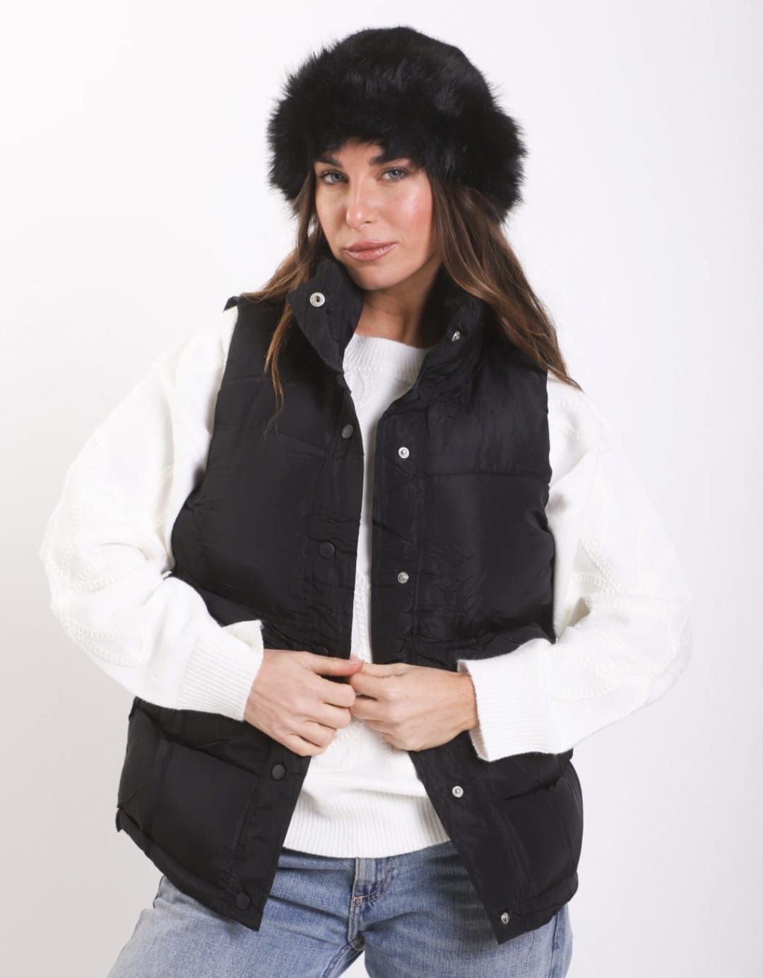 Mujer con chaleco negro y gorro de piel sobre una camisa blanca, de pie frente a un fondo blanco.