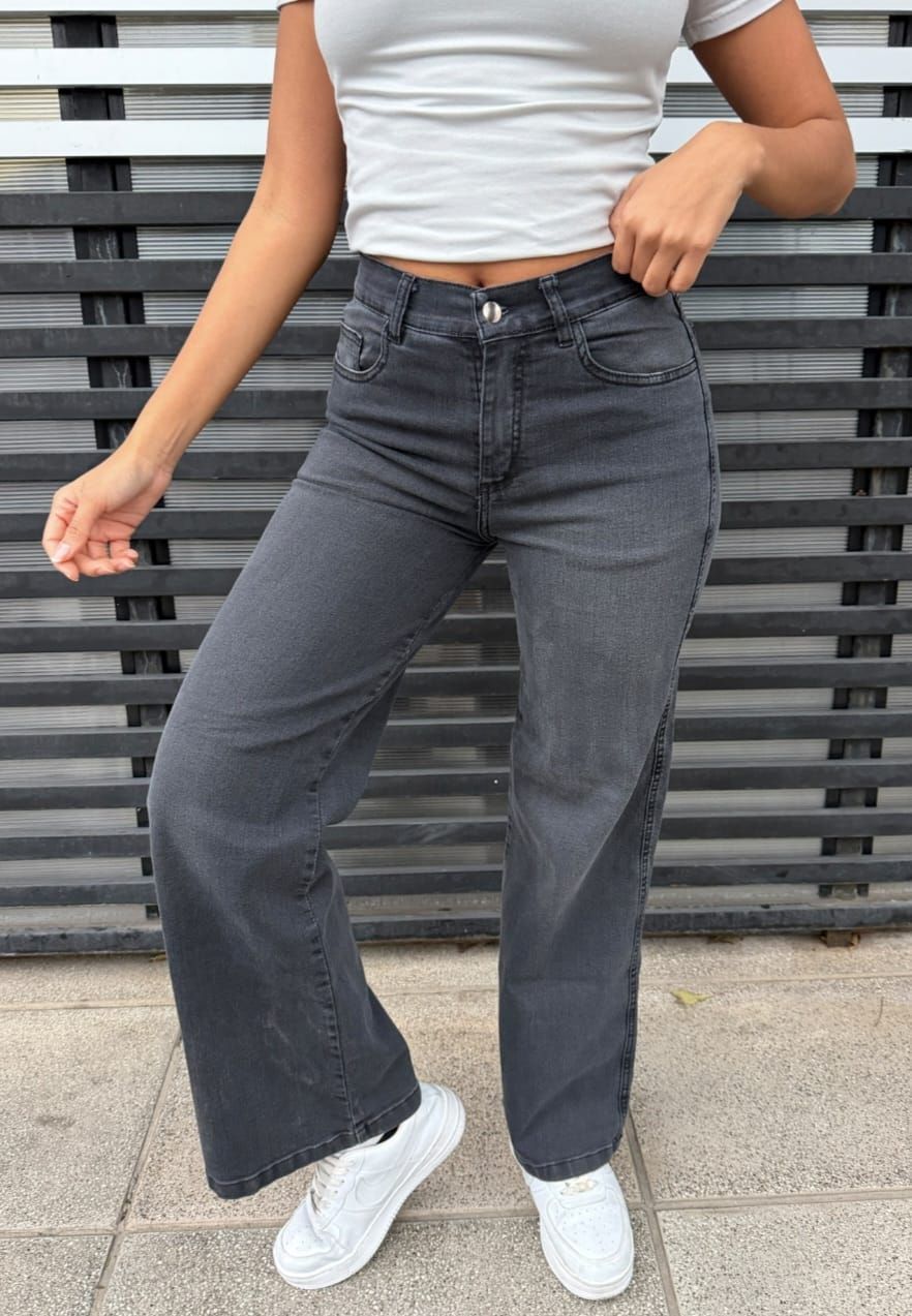 Una persona vestida con una camiseta blanca y vaqueros oscuros, con zapatillas blancas, está de pie frente a una pared gris de listones.