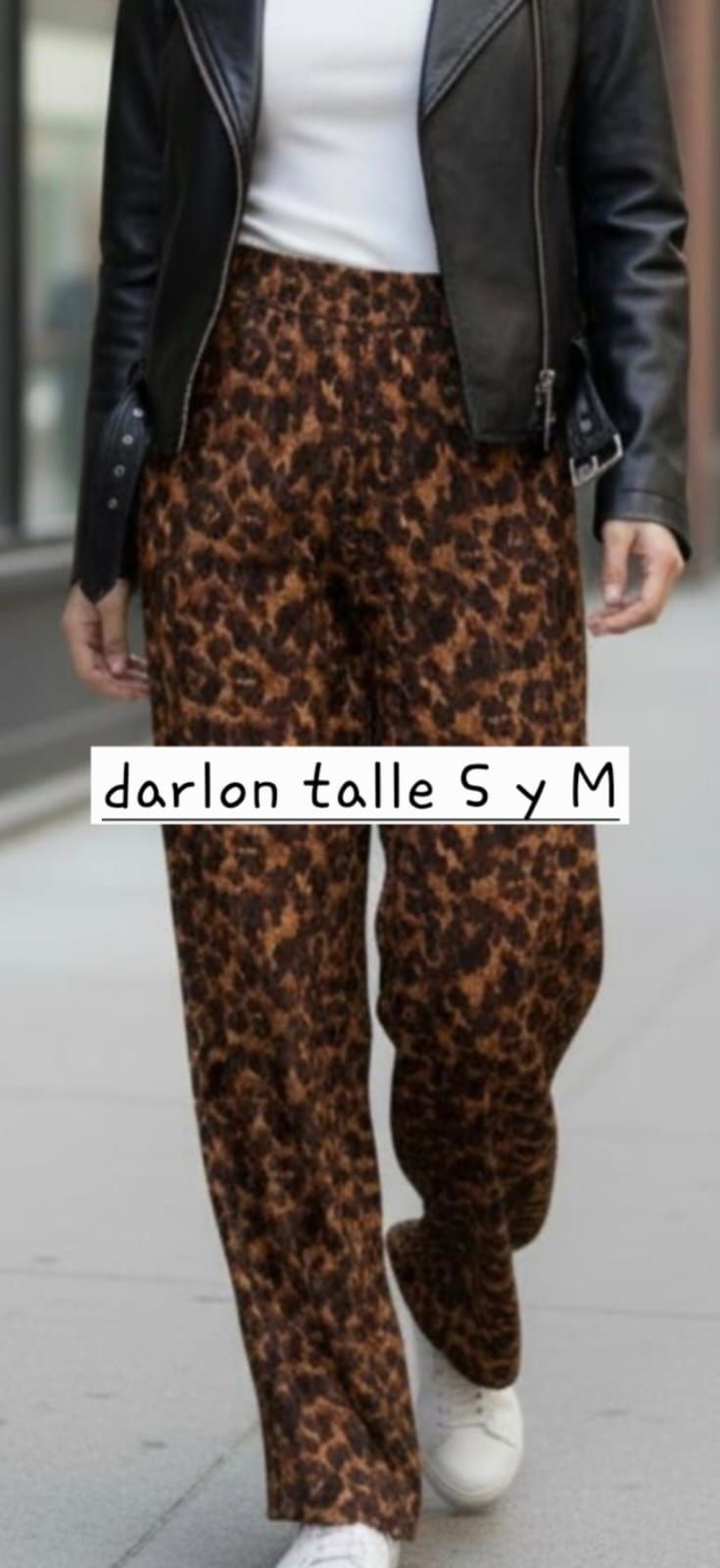 Persona caminando por una acera de la ciudad con pantalones estampados de leopardo, chaqueta negra y zapatillas blancas.