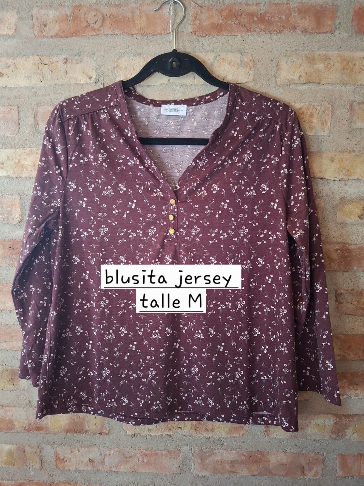 Blusa granate con estampado floral colgada en una percha contra una pared de ladrillos.