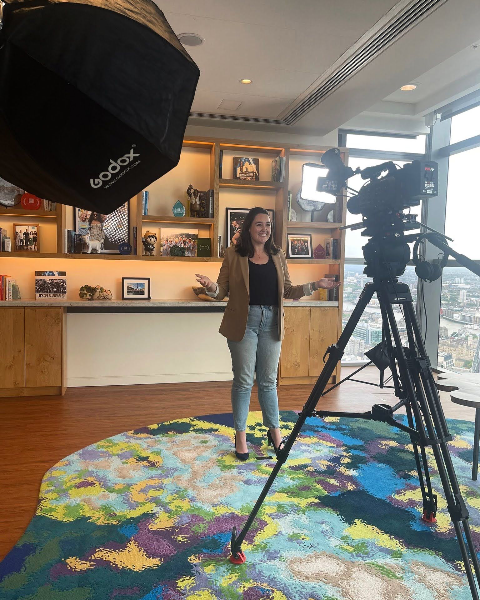 Woman filming video in a room with city view, standing near a camera and softbox light.