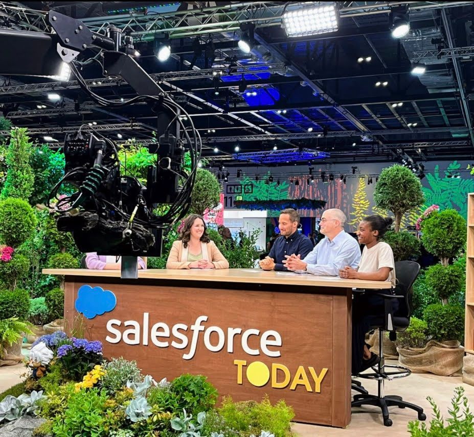 Salesforce Today set with four people at a desk. Lush greenery surrounds them. Studio lights overhead.